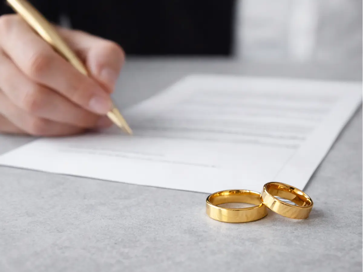 Regimes de casamento  representada por pessoa assinando um documento enquanto duas alianças estão apoiadas ao lado.