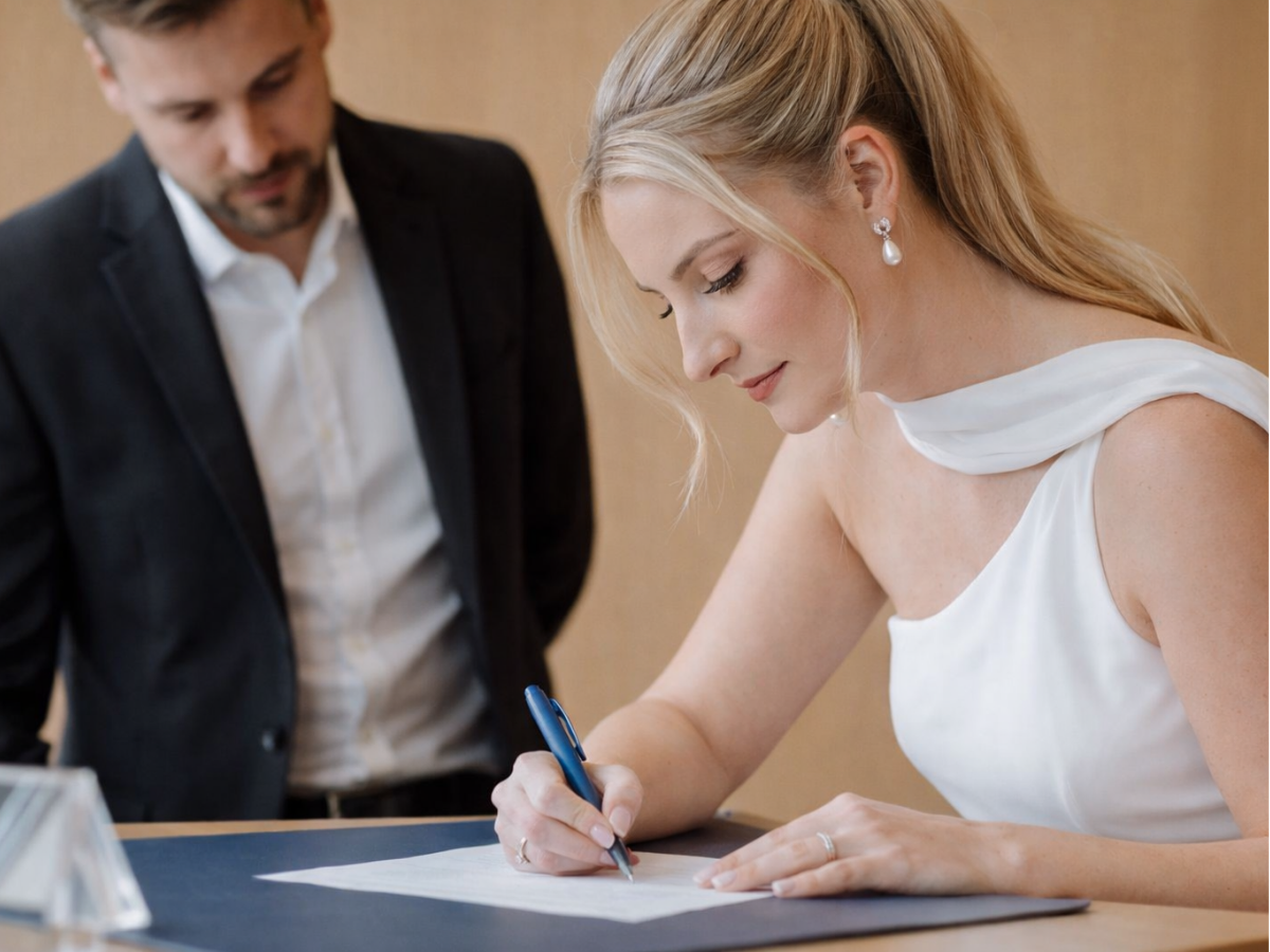 Mulher assinando um documento com outra pessoa ao lado.