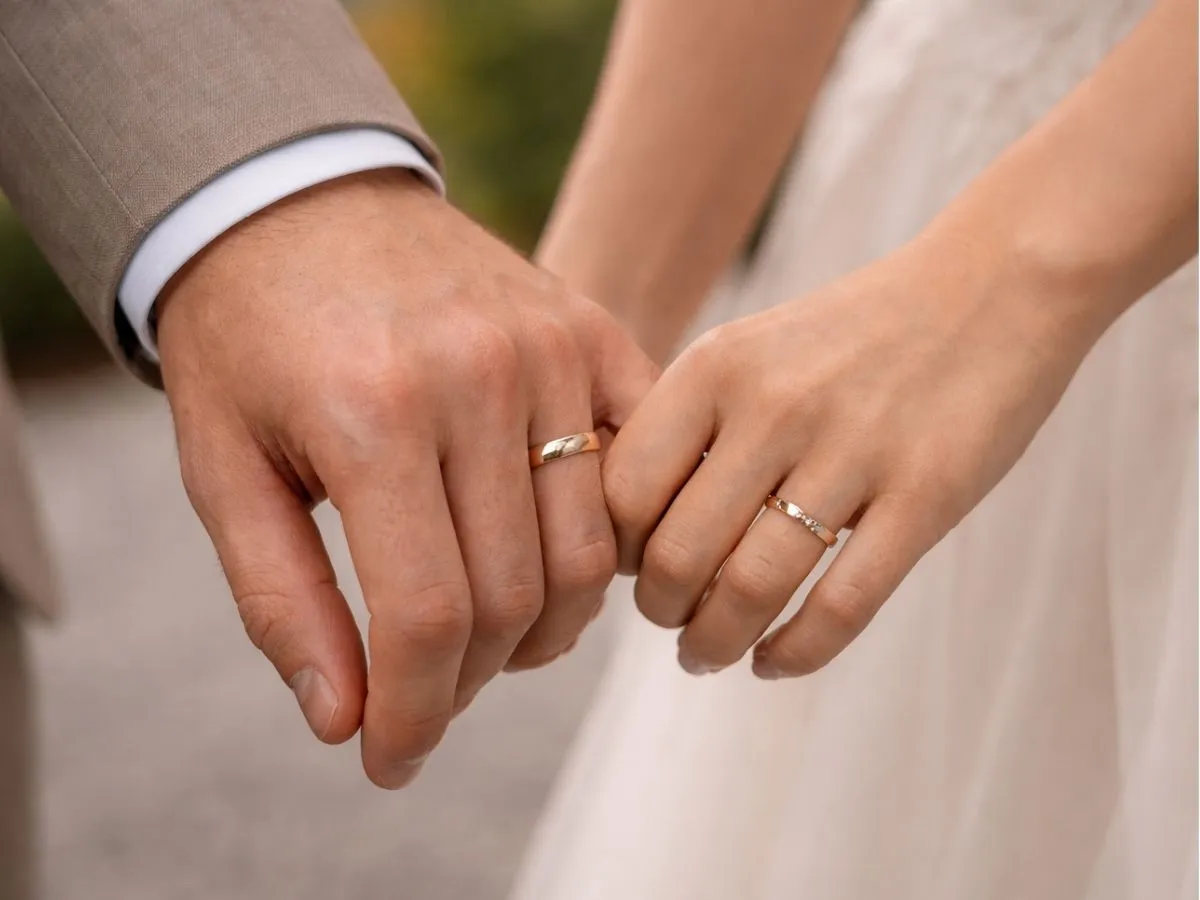 Mãos de um casal lado a lado usando anéis simples em tom dourado.