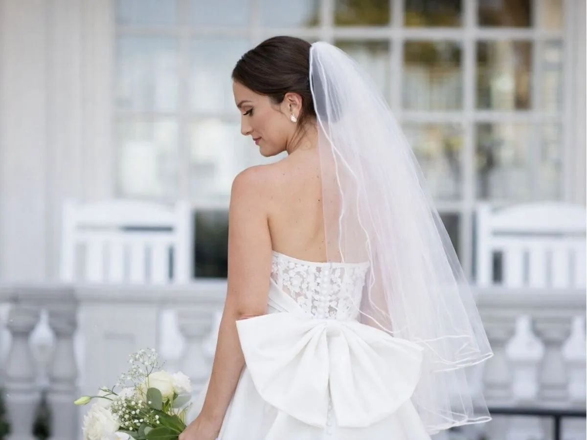 Noiva vista de costas usando vestido branco com laço e véu longo.