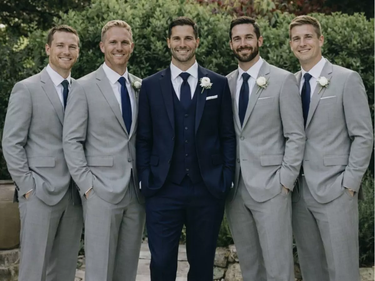 Noivo em terno azul-marinho ao centro, acompanhado por padrinhos em ternos cinza claro em ambiente externo.