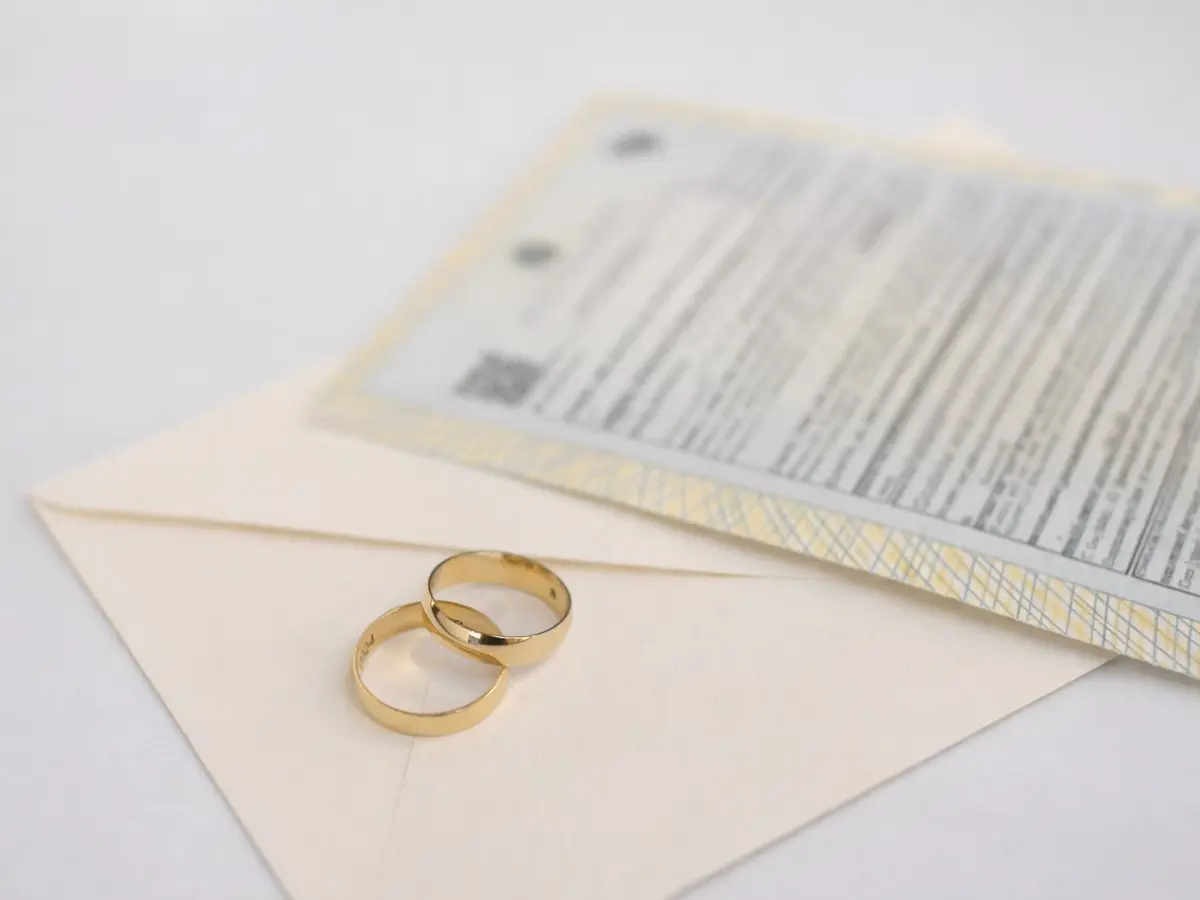 Alianças de casamento douradas sobre um envelope claro, com a certidão de casamento ao fundo.