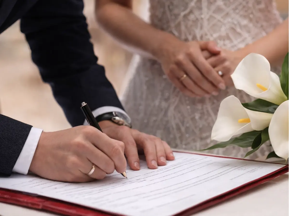 Noivo assinando o registro de casamento civil enquanto a noiva acompanha, com buquê de flores brancas ao lado.