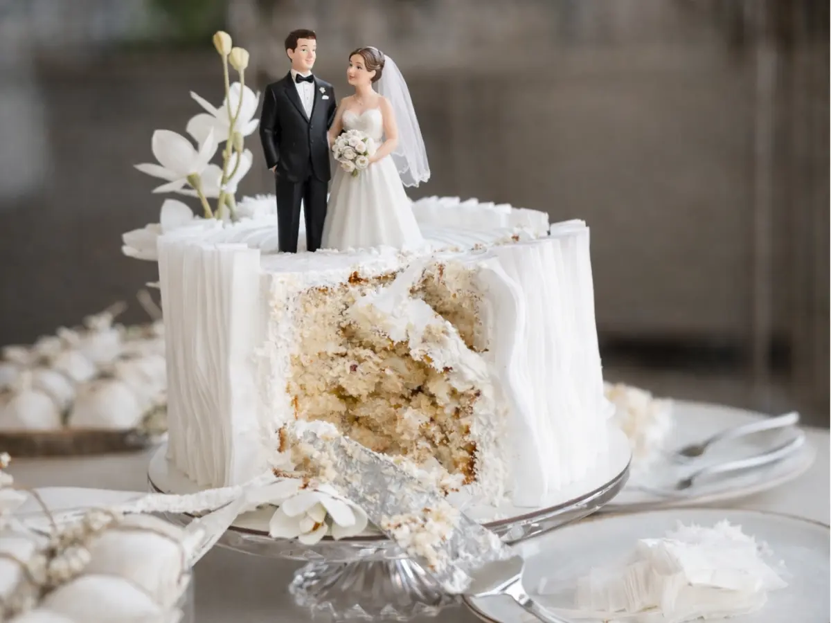Bolo de casamento branco com topo de noivos, fatia cortada revelando o interior e espátula apoiada no prato.