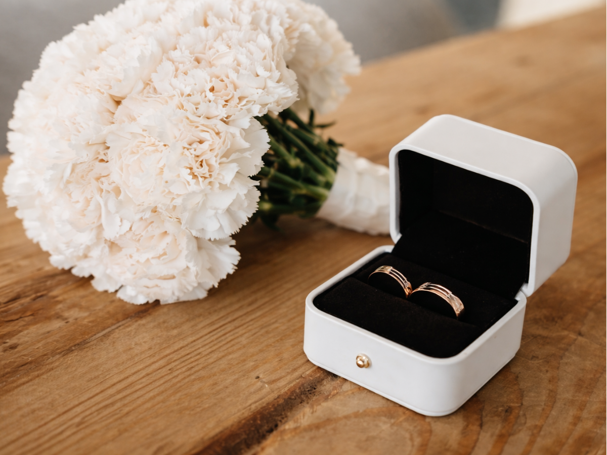 Buquê branco ao lado de estojo branco com anel de noivado sobre mesa de madeira.