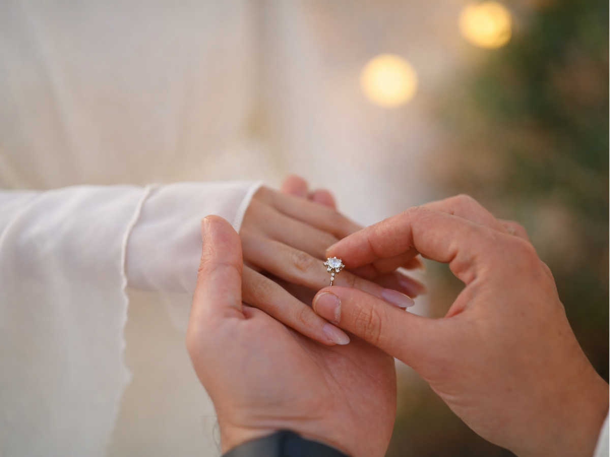 Mãos colocando anel de noivado no dedo da noiva em ambiente romântico.