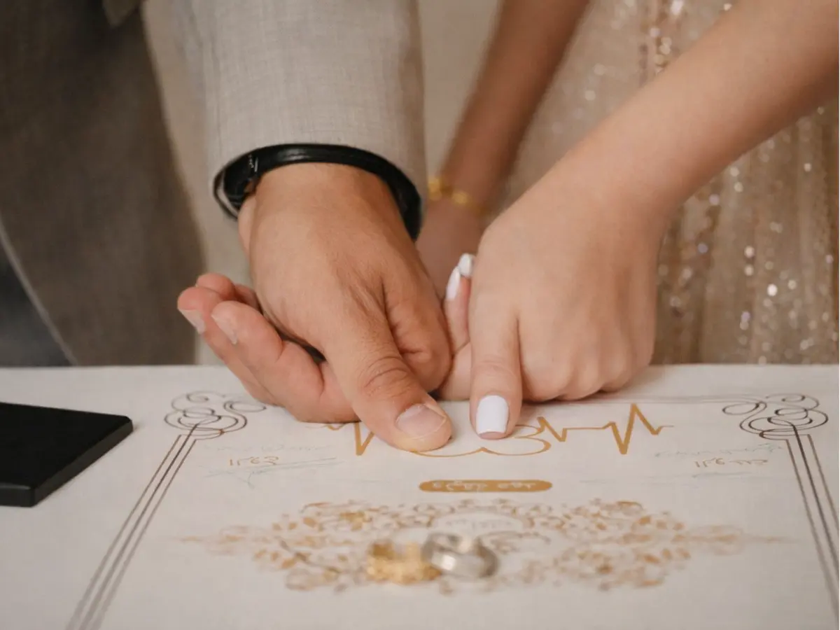 Mãos de um casal apontando a assinatura na certidão de casamento, com alianças sobre o documento.